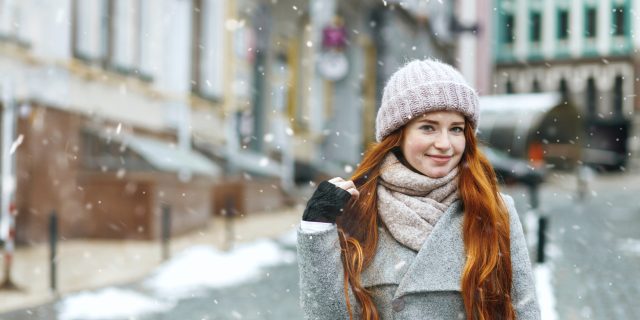 A young red-haired woman wearing a gray winter coat poses confidently as snow falls around her.