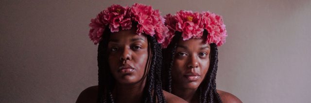 Two Black women with flower crowns against a brown backdrop stare into the camera