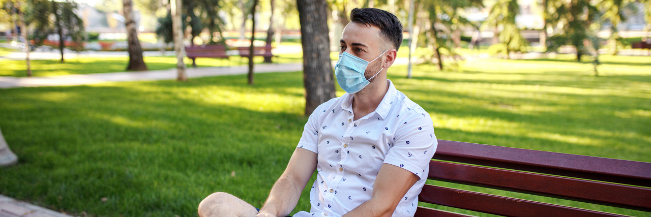 Ways to Improve Your Mental Health During the COVID-19 Pandemic A man with dark brown hair and dark brown eyes wearing a white shirt with black polka dots and a pair of blue shorts sits on a park bench while wearing a mask.