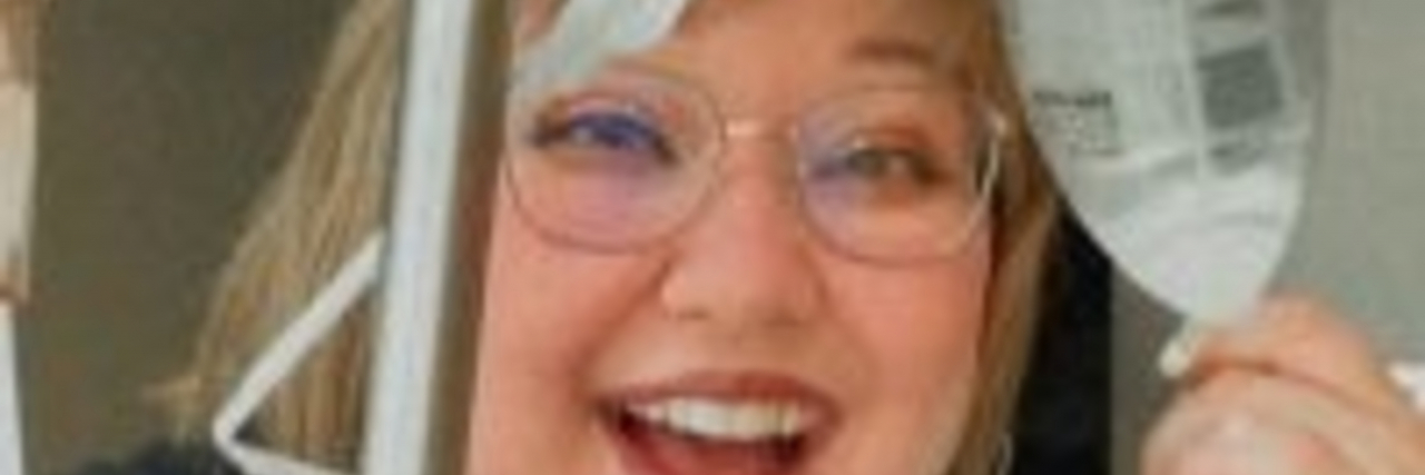 Seeing the Future With Curiosity, Not Hope With a Progressive Illness Megan, a woman with short blonde hair and glasses wearing a black T-shirt, smiles cheerily while holding an IV bag.