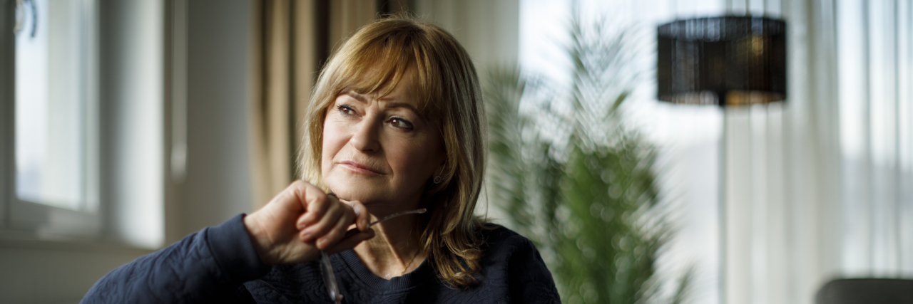 When Dementia Slowly Takes Your Loved One Away From You A woman with red shoulder-length hair wearing a navy blue sweater looks pensive while sitting on a couch and holding a pair of glasses.