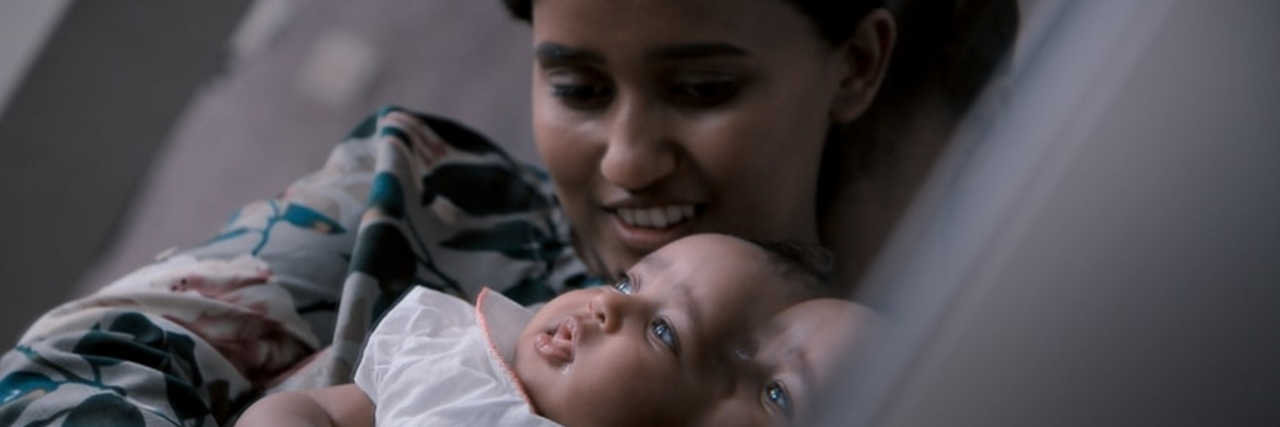 A white woman with brown eyes and brown hair tied back holds a baby wearing white.