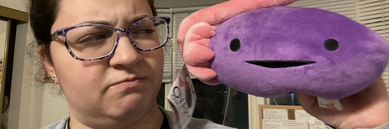 Anxiety and Dismissive Doctors Made Me Doubt My Chronic Pain Is Real photo of the author, woman holding a smiling plush of an ovary and looking confused