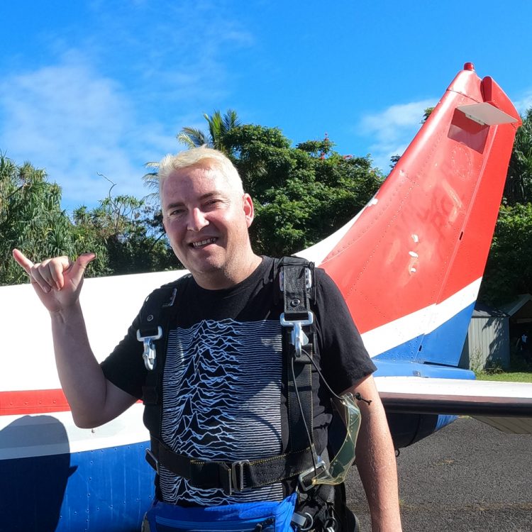 A white man throws a shaka hand sign outside of a plane. He's wearing a harness, prepped to bungee jump. His shirt is black, and the plane has red and blue stripes.