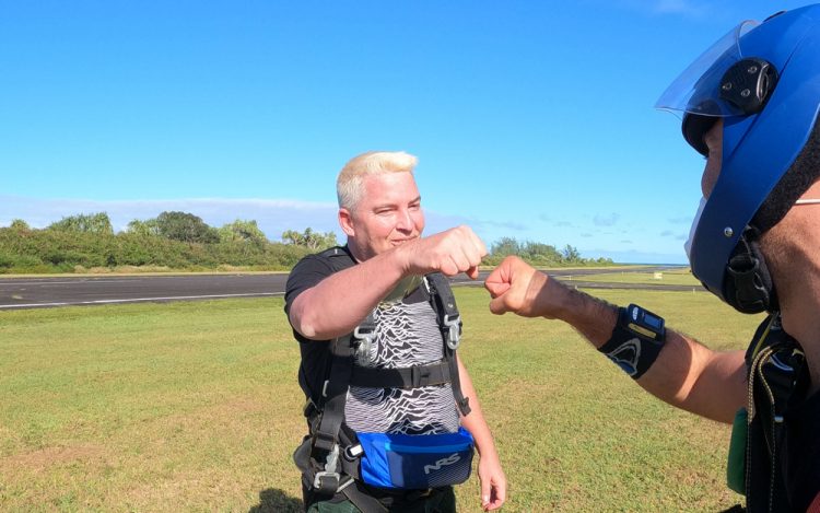 The two men fist bump. They wear all black and are on the ground, green grass below them and blue sky above them.