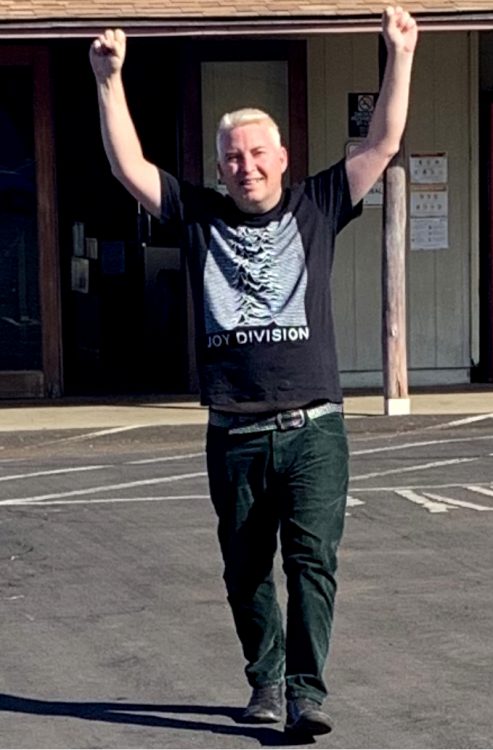 The man raises his fists high in the air, triumphant! He is still in a Black shirt and black pants. I think it's a joy division shirt?