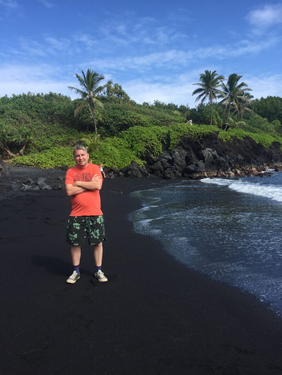 A man stands on a black sand beach, in a bright red shirt with his hands crossed. The sky is blue above him and there's greenery behind him.