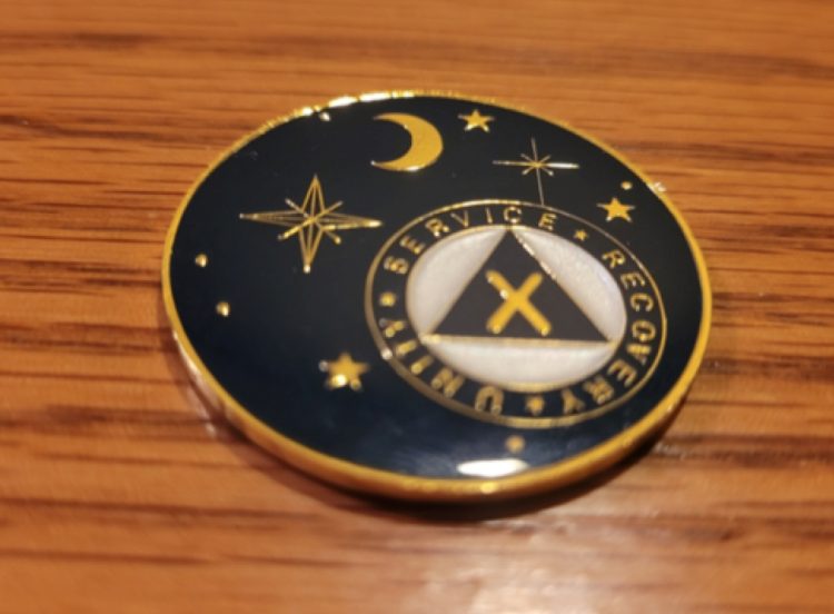 A black sobriety coin that has gold stars and moons on it. There's a white circle with a black triangle and a gold x in the center.