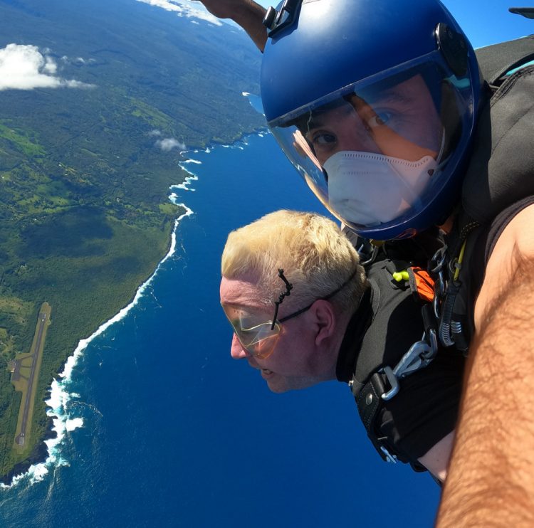 Two men are attached to one another jumping out of an airplane. One man is definitely scared out of his life, the other looks up at the camera. Blue ocean and green forrest is below them