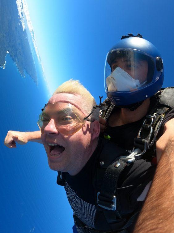 A man has his mouth open, smiling as he jumps out of the plane with goggles on. The man above him has a helmet on and a mask. The helmet is blue. The blue ocean is blow them