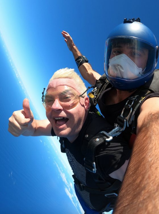 The man throws a thumbs up at the camera, still free falling. Blue sky and ocean surround the two men. The man on top has a mask and helmet on. 