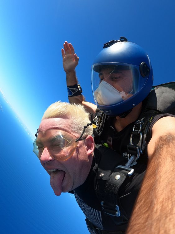 Now, the man is sticking out his tongue. Outside of that, literally nothing has changed from the earlier two images. They're still suspended in air, the man on top has a blue helmet on and a mask. They're surrounded by blue skies and ocean. 