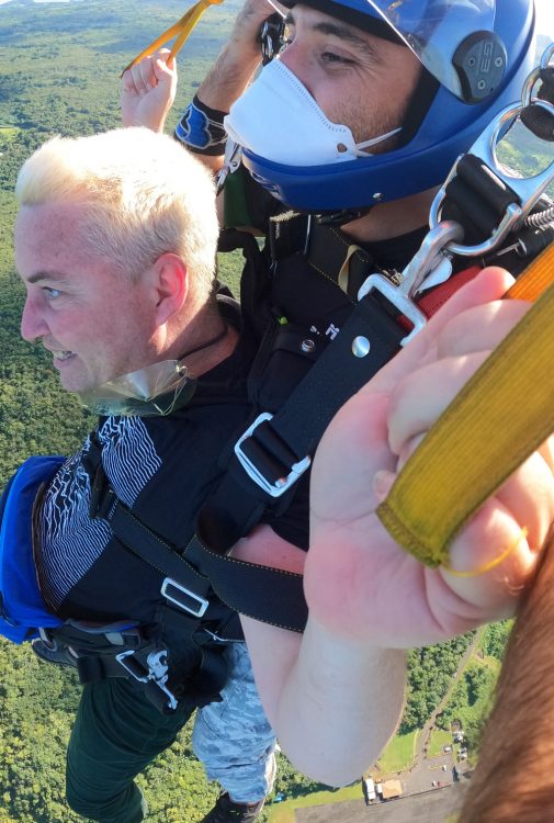 The man on the bottom grits his teeth as they descend. The camera is angled above them, with his fist closest to the camera. Under them are green trees