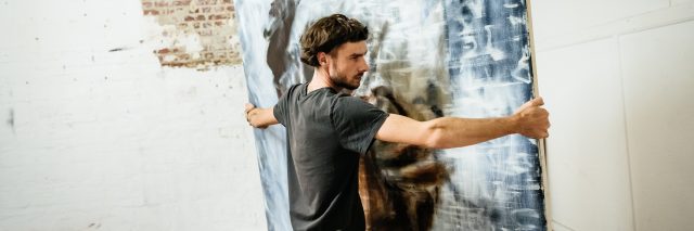 A brunette man wearing a gray T-shirt and jeans carries a painted canvas in a warehouse with a brick wall.