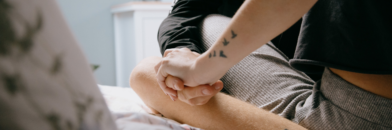 intimate photo of two people sitting on a bed with legs crossed and holding hands