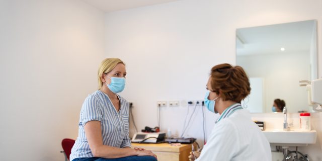 A blonde woman wearing jeans and a blue-and-white striped shirt as well as a mask speaks with a female doctor with short, curly red hair. The doctor is also wearing a mask.