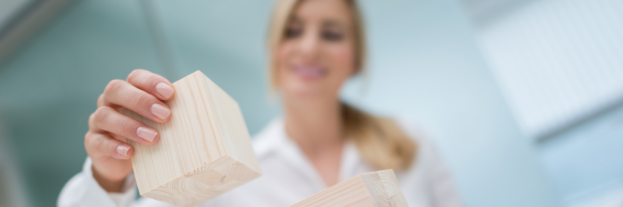 Why I View My Recovery Progress as 'Building Blocks' After My Suicide Attempt Hospitalization A blonde, long-haired woman wearing a white shirt stacks wooden blocks on top of each other.