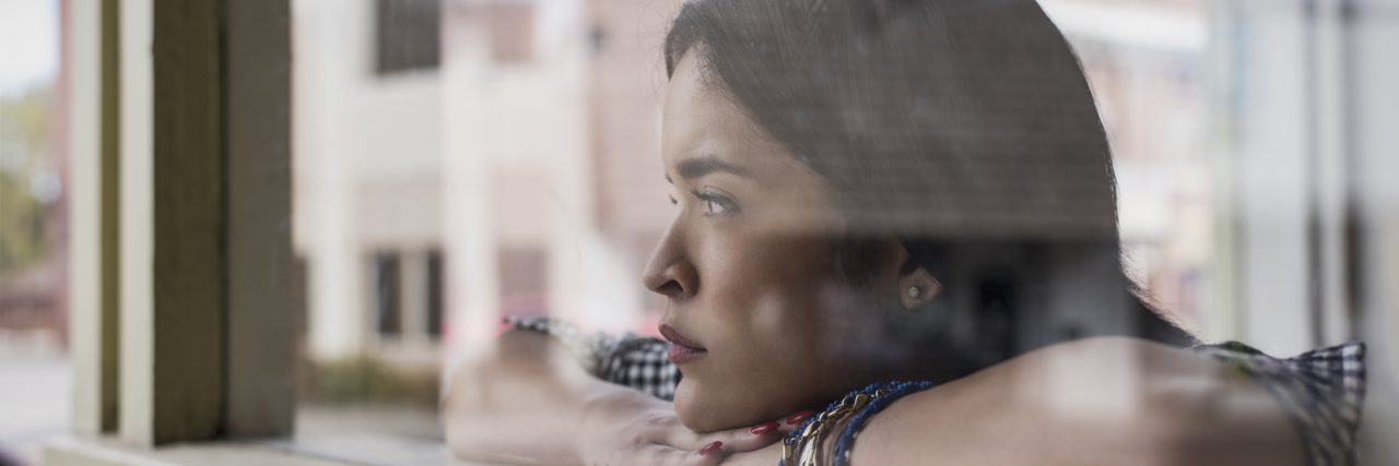 A woman of color perches her arms on a windowsill and looks out the window.