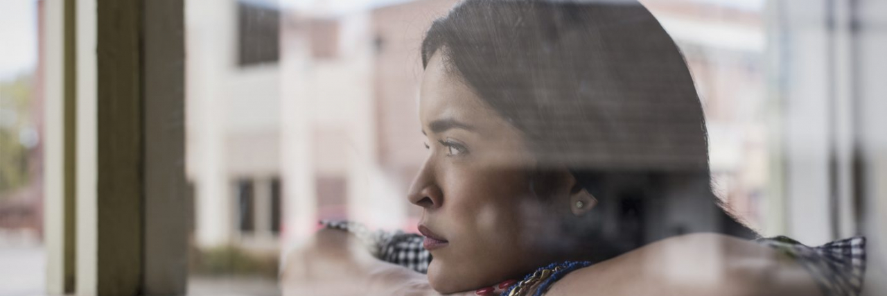 A woman of color perches her arms on a windowsill and looks out the window.