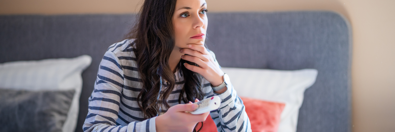 A woman with long brown hair and brown eyes sits on a bed while wearing a striped long-sleeved shirt and jeans. She is holding a remote control.