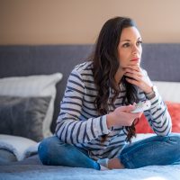 A woman with long brown hair and brown eyes sits on a bed while wearing a striped long-sleeved shirt and jeans. She is holding a remote control.