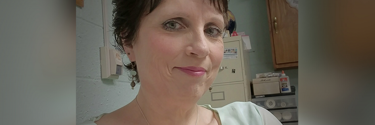 What Helps a Woman in Her Fifties With Post-traumatic Stress Disorder A woman with short brown hair and blue eyes wearing a white blouse and a necklace smiles softly for the camera.