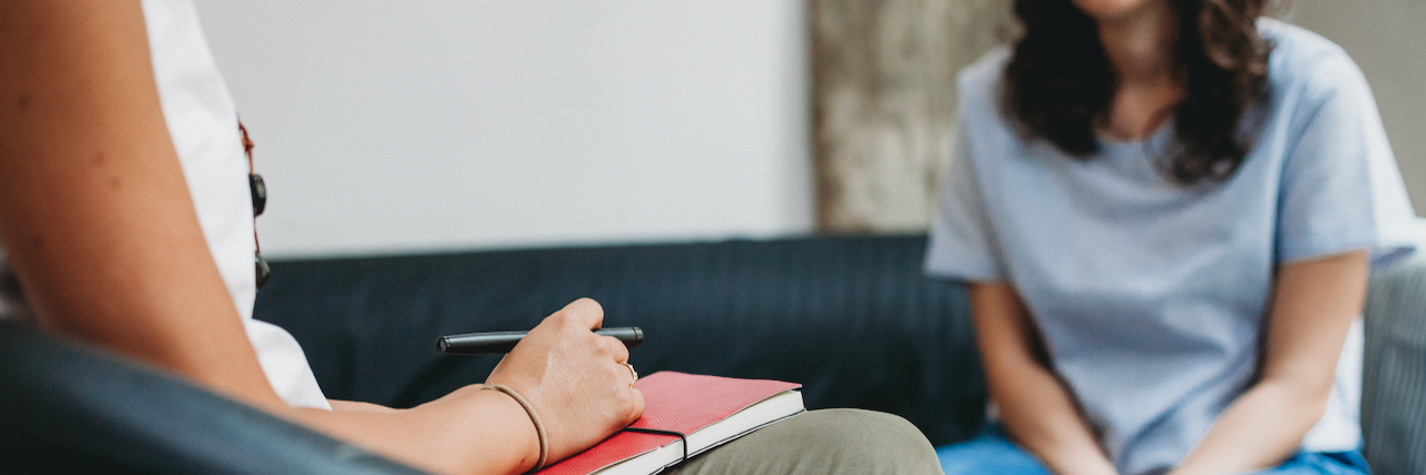 What Self-Disclosure Can Mean in a Therapeutic Relationship Woman sitting on couch speaking with her therapist, who is holding a red notebook and pen