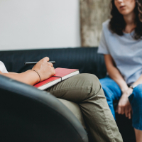 Woman sitting on couch speaking with her therapist, who is holding a red notebook and pen