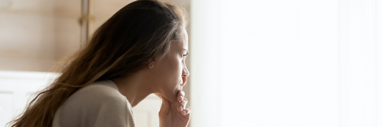 Mourning Emotional and Physical Losses With Chronic Illness Woman with long, brown hair sitting on bed and looking out a bright window
