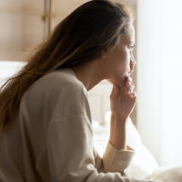 Woman with long, brown hair sitting on bed and looking out a bright window