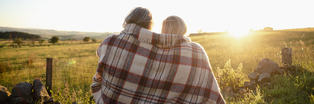 A couple sitting on a field with a blanket wrapped around them, sun shining