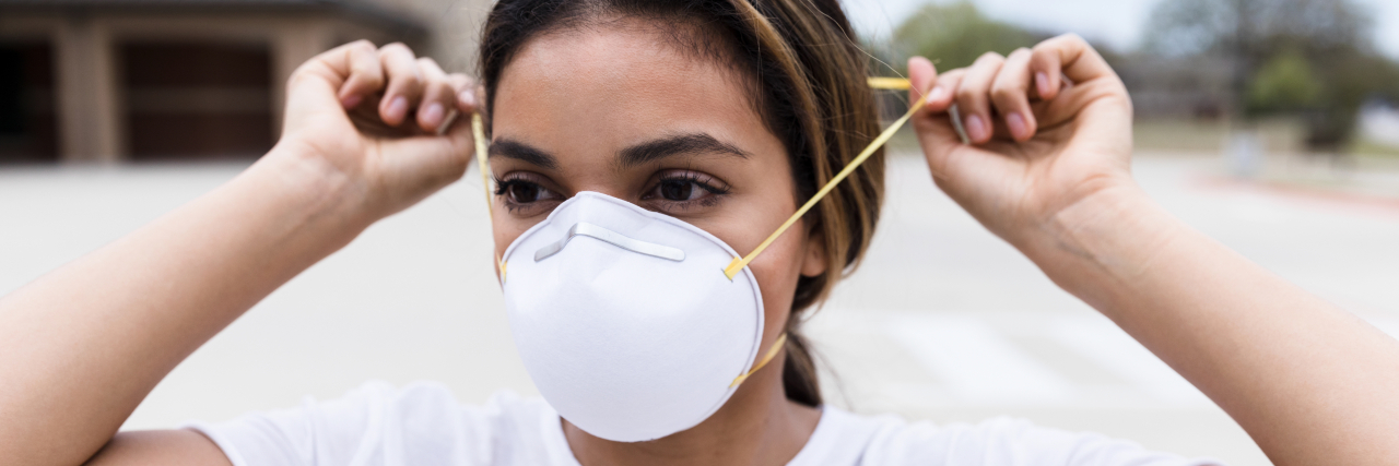 How Being 'Done' With COVID Safety Hurts Those With Chronic Illness Woman standing outside places an N95 mask on her face for protection during the pandemic.