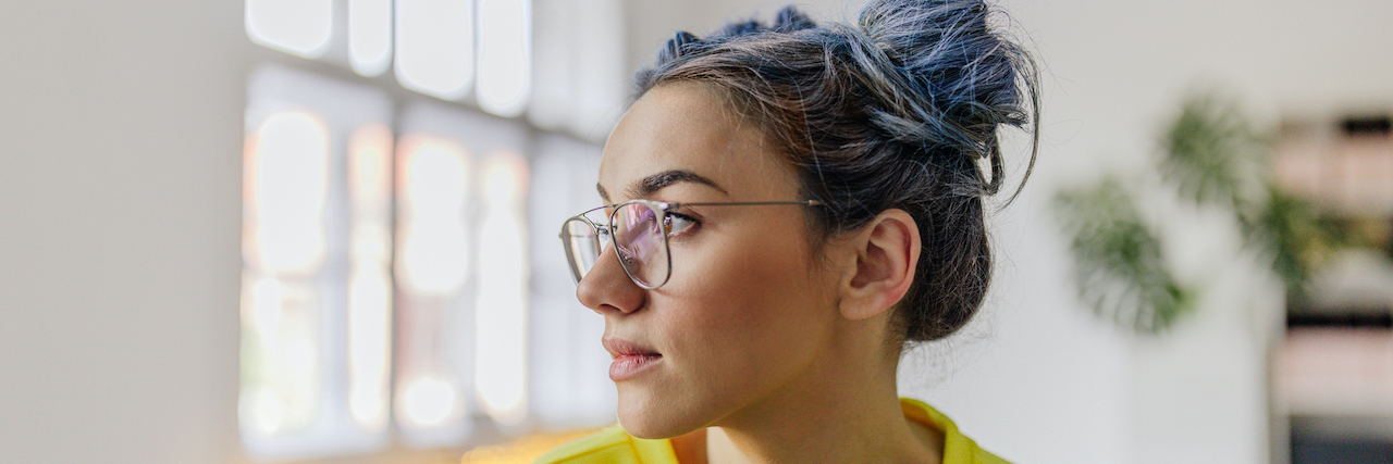 Being Open About My Chronic Pain Helps Me Cope With It Woman with blue on the bottom half of of her hair, wearing glasses and looking to the side