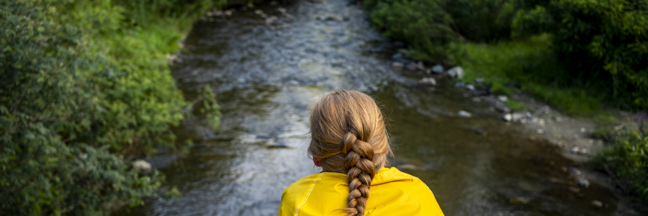Why I Struggled to Accept That I'm Disabled By Dercum's Disease Woman in a yellow jacket is sitting on a bridge over the river.