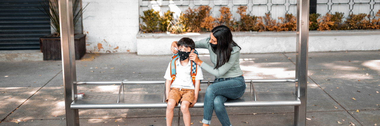 Mother and son at the bus station, she is helping him with a mask.
