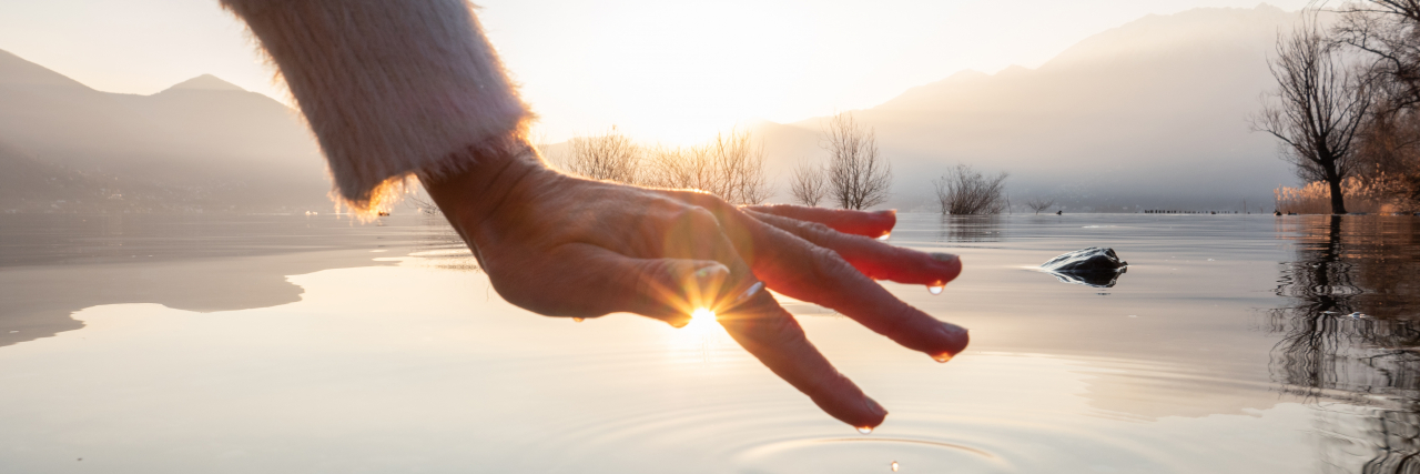 The Great Teacher That Is Chronic Illness Detail of hand touching water surface of lake at sunset