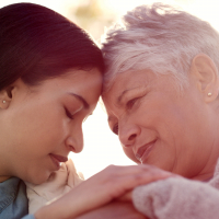 An adult daughter and mother. Their heads are pressed against each other and they are holding hands.