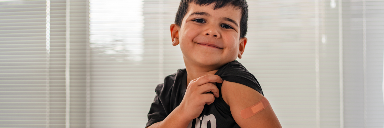 How Parents Can Make Educated Choices About the COVID Vaccine for Kids Child smiling, showing bandage on shoulder after receiving the COVID vaccine.