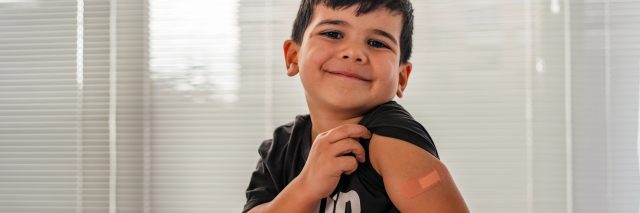 Child smiling, showing bandage on shoulder after receiving the COVID vaccine.