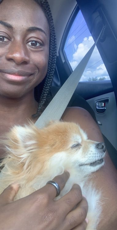 A very cute but very distraught Black girl holds an orange sable pomeranian dog. The dog's eyes are closed, and she's in a car. The girl tries to smile for the camear, but has tears in her eyes. She has braids. The sun is shining bright outside.