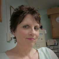 A woman with short brown hair and blue eyes wearing a white blouse and a necklace smiles softly for the camera.