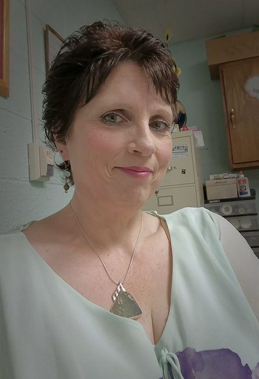 A woman with short brown hair and blue eyes wearing a white blouse and a necklace smiles softly for the camera.