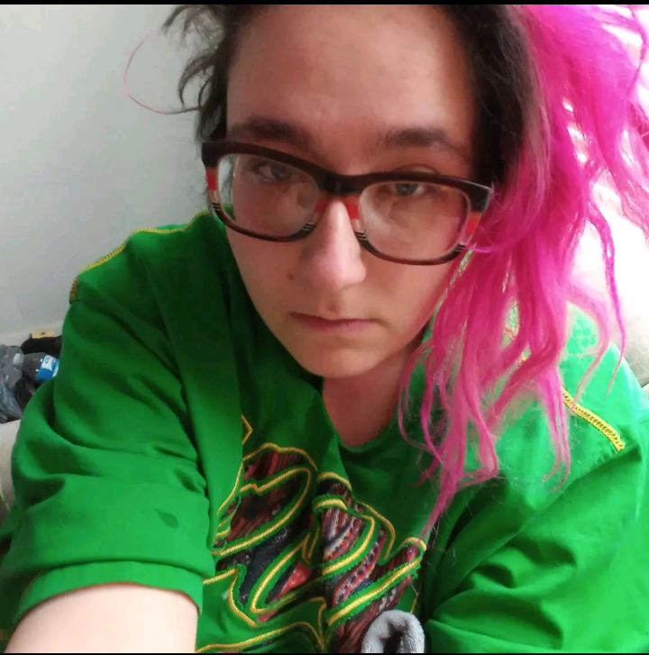 A person with bright pink hair in a green shirt stares forlornly at the camera. They wear glasses and are against a wall.