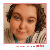 In a light red frame is a person with blue eyes and brown curly hair. In red writing below is "The In-Between by The Mighty"
