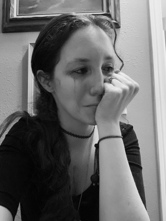 In black and white, a woman has tear stains on her face. She wears a dark black shirt