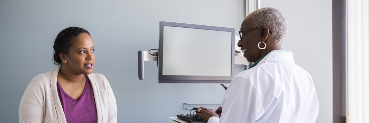 Coping With Doctors Telling You You're 'Fine' With Chronic Illness A black woman wearing a cream-colored cardigan, a purple top, and jeans speaks with a black woman doctor while sitting on an exam table.