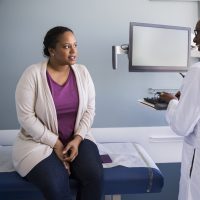 A black woman wearing a cream-colored cardigan, a purple top, and jeans speaks with a black woman doctor while sitting on an exam table.