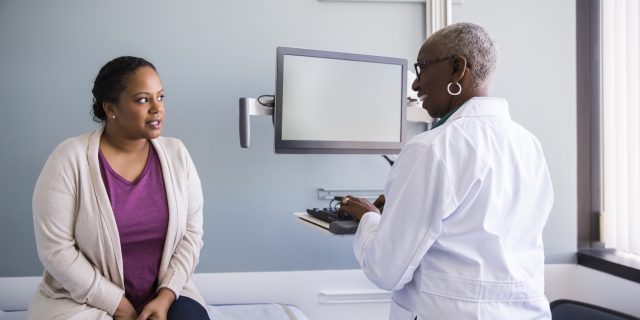 A black woman wearing a cream-colored cardigan, a purple top, and jeans speaks with a black woman doctor while sitting on an exam table.