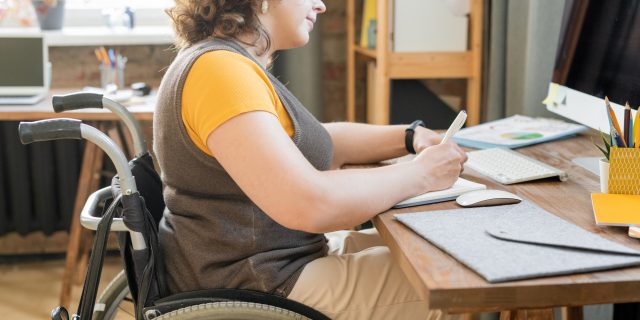 Close-up photo of a woman wearing a gray vest, an orange shirt, and khaki pants writing while she sits in a wheelchair.