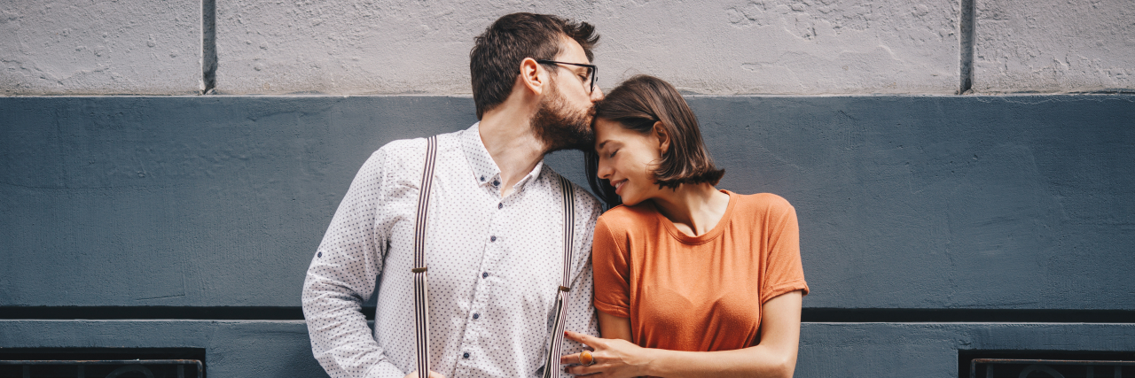 Finding Love and Marriage as a Woman With a Learning Disability A white man wearing a white button-down shirt, blue jeans, and suspenders holds hands with and kisses a white woman with short brown hair wearing an orange top and a gray skirt.